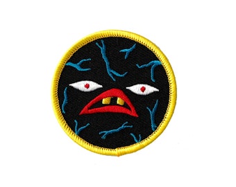 May include: A black and yellow embroidered patch with a cartoon face. The face has red eyes, a red mouth, and two white teeth. The background is black with blue veins.