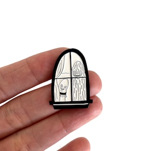 Puede incluir: Un pin de esmalte en blanco y negro con una ilustración de una ventana. El diseño incluye una cortina, una planta y una criatura sonriente. El pin se sostiene en una mano.