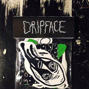 May include: A clear plastic bag with a black header that says "DRIPFACE" in white lettering. The bag contains a variety of black and white stickers with a green leaf sticker. The stickers feature a black and white illustration of a face with dripping eyes.