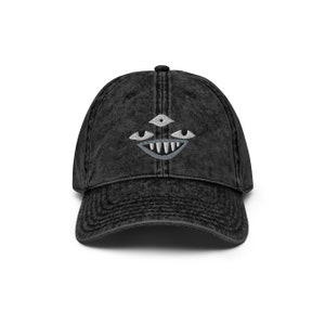 May include: A black vintage washed denim baseball cap with a white embroidered design of a face with three eyes and a toothy grin.