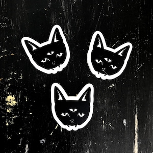 May include: Three black cat stickers with white outlines. Each cat has three eyes and a slightly sinister expression.