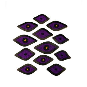 May include: A collection of eleven embroidered patches in a diamond eye shape. Each patch features a purple center with a yellow iris and a black border. The patches are arranged on a white background.