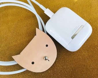 May include: A white phone charger with a leather cat-shaped cable holder on a brown surface.