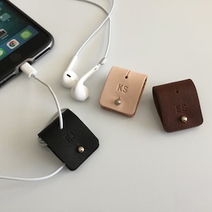 May include: Three leather cord organisers with initials 'EJ', 'KS', and 'ES' embossed on them. The organisers are brown, black, and beige.