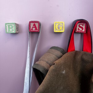May include: Three wooden blocks spelling "BAGS" are mounted on a wall. A brown canvas bag hangs from the "S" block.