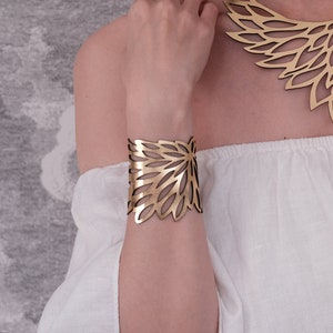 May include: A gold-colored statement necklace and matching cuff bracelet. The jewelry features a floral design with intricate cut-out patterns. The necklace sits on a black cord, and the cuff is worn on the wrist. The jewelry is displayed against a neutral background.