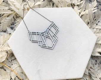 May include: A silver geometric necklace with a modern design. The necklace is displayed on a white marble surface.