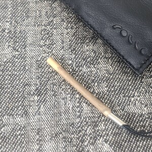 May include: A black leather case with the embossed word "rocco" and a small, cylindrical object with a silver tip. The object is light brown and tan. The items are on a textured gray and white surface.