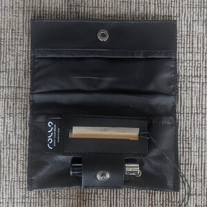 May include: Black leather pouch with a snap closure, open to reveal a compartment with a small pack of rolling papers and a lighter. The pouch is designed to hold smoking accessories.