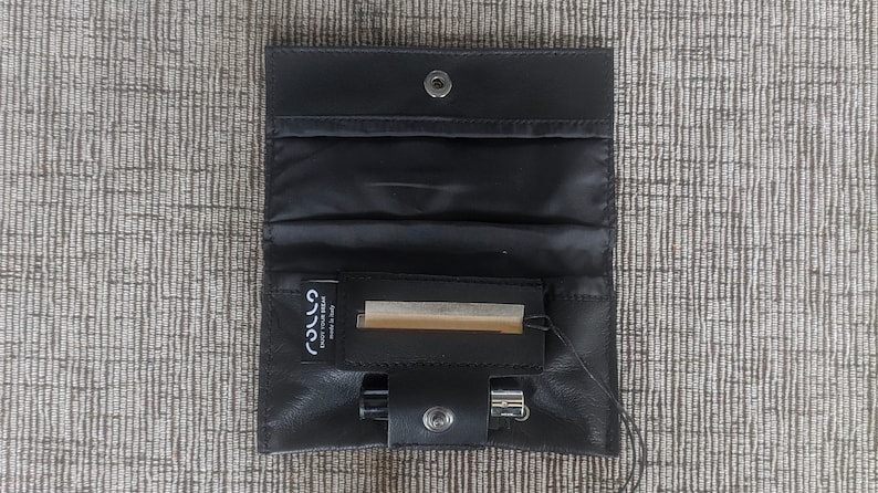 May include: Black leather pouch with a snap closure, open to reveal a compartment with a small pack of rolling papers and a lighter. The pouch is designed to hold smoking accessories.