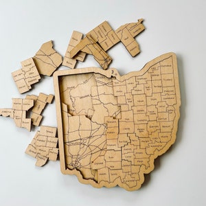 May include: Wooden Ohio state puzzle with individual county pieces. The puzzle is made of light-colored wood, with each county labeled. Some pieces are removed, showing the map's design and the county names.