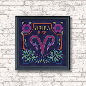 May include: A framed cross-stitch design featuring the astrological sign Aries, with the words "Aries Fire" in a banner above the ram symbol. The design is in a dark blue background with pink, green, and yellow accents.