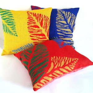 May include: Four square cushions in vibrant colours. One yellow cushion with green and red leaf design, one red cushion with green and gold leaf design, and two blue cushions with gold leaf design. The cushions are made of woven fabric.