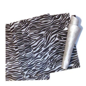 May include: A stack of rectangular sheets featuring a black and white zebra stripe pattern. The top sheet is partially rolled at the right edge, revealing a smooth, light gray underside. The sheets are arranged in a slightly overlapping, layered formation.