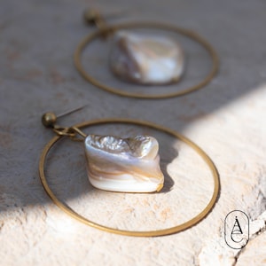 Puede incluir: Un par de pendientes de aro dorados con un gran colgante de concha blanca iridiscente de forma irregular.