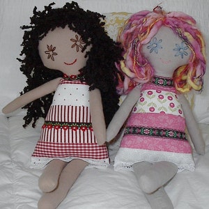 May include: Two handmade fabric dolls with yarn hair. The doll on the left has brown yarn hair and a red and white gingham dress. The doll on the right has pink and yellow yarn hair and a pink and white floral dress.