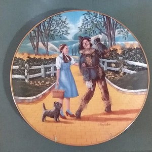 May include: A round ceramic plate with a colorful image of Dorothy from the Wizard of Oz walking down the yellow brick road with the Scarecrow. The plate has a gold rim and a yellow background.