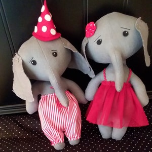 Puede incluir: Dos peluches de elefante de tela gris, uno lleva un atuendo a rayas rojas y blancas y un sombrero de lunares rojos y blancos, el otro lleva un vestido rojo con una flor roja.