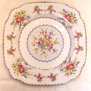 May include: White square plate with a floral cross-stitch design. The plate features a central bouquet and floral clusters around the rim, with pink, yellow, and blue flowers. A blue zig-zag border frames the design.