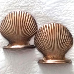 May include: A pair of metallic bronze-toned bookends shaped like large, fanned scallop shells. Each bookend features a semi-circular base and a textured surface with radiating vertical ridges that extend from the bottom to the rounded top edge.