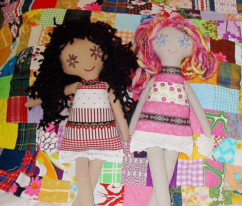May include: Two handmade rag dolls with yarn hair. One doll has brown hair and wears a red and white striped dress with white polka dots. The other doll has pink and yellow yarn hair and wears a pink and white floral dress. The dolls are sitting on a colorful patchwork quilt.