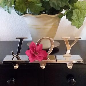 May include: A set of three silver-toned metal stocking holders spelling "JOY". A red poinsettia flower is placed between the "O" and the "J". The holders are on a black surface, with a decorative plant in the background.