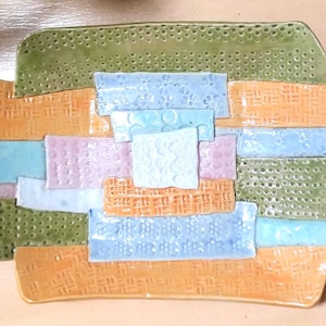 May include: A colorful ceramic art piece with a patchwork design. The piece features rectangular and square shapes in shades of orange, green, blue, and pink. The textures vary, with some areas having a dotted or woven pattern.