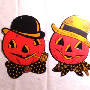 May include: Two flat cardboard Halloween decorations featuring orange jack-o'-lantern faces. One wears a black bowler hat and a black polka-dot bow tie with a pipe. The other wears a yellow hat and a black patterned neckerchief with a pipe.