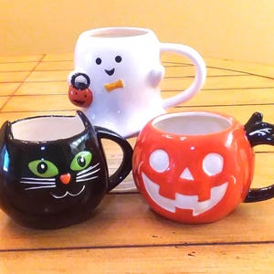 May include: Three ceramic mugs featuring Halloween designs. One white ghost mug holding a small orange pumpkin bucket, one black cat mug with green eyes and white whiskers, and one orange jack-o'-lantern mug with a carved face and black handle.