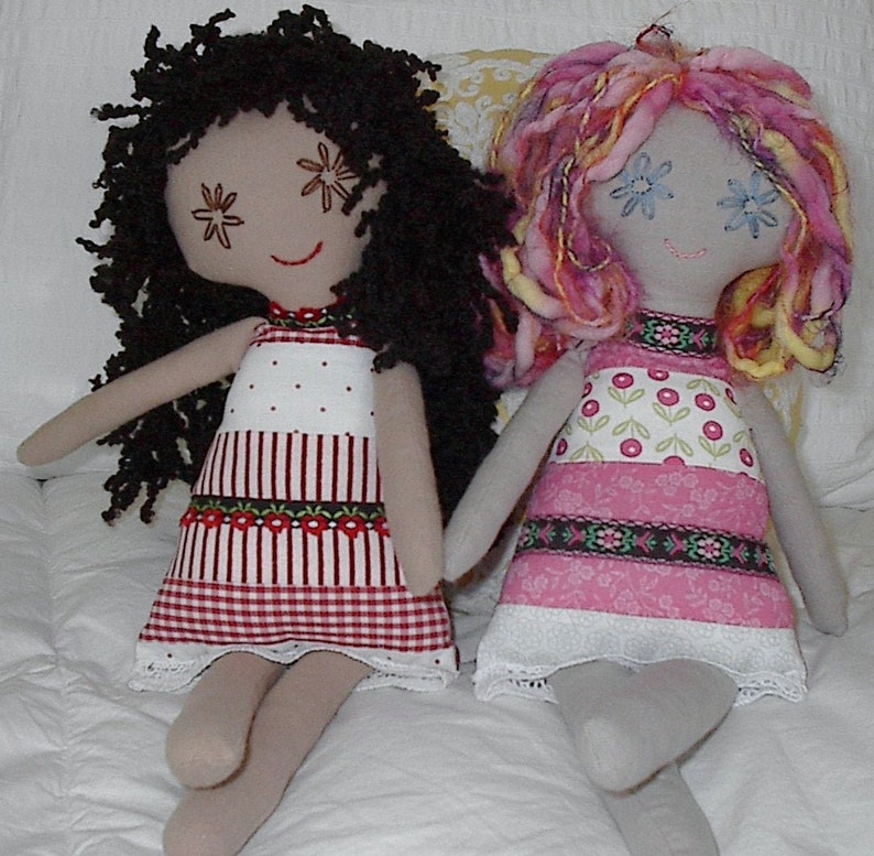 May include: Two handmade fabric dolls with yarn hair. The doll on the left has brown yarn hair and a red and white gingham dress. The doll on the right has pink and yellow yarn hair and a pink and white floral dress.