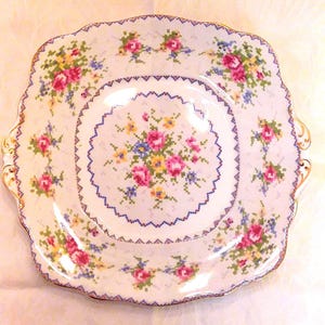 May include: Square, off-white porcelain plate with a floral cross-stitch design. The plate features pink, yellow, and blue flowers with a blue zigzag border and gold trim. A smaller plate sits on top.