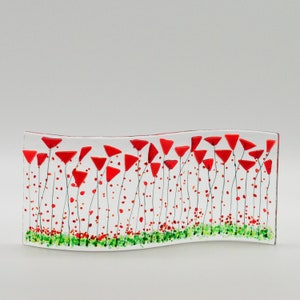 Może przedstawiać: Szklana praca artystyczna przedstawiająca pole czerwonych maków. Maki są ułożone w zakrzywioną linię na białym tle. Praca wykonana jest ze szkła i ma fakturowaną powierzchnię.