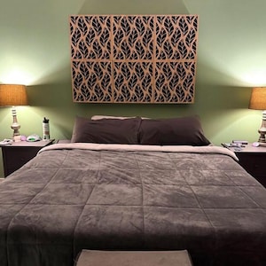 May include: A bedroom scene featuring a bed with a gray comforter and pillows. Above the bed is a decorative wall panel with a leaf design in black and tan. Two lamps flank the bed, casting a warm glow.