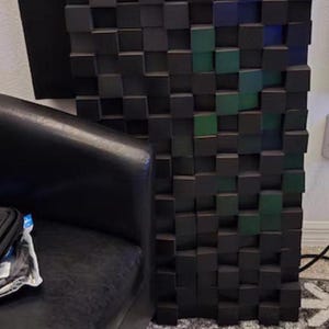 May include: A textured wall panel made of small, square blocks in shades of black, grey, blue, and green. The panel is next to a black leather armchair, with a patterned rug below. The panel is likely for soundproofing.