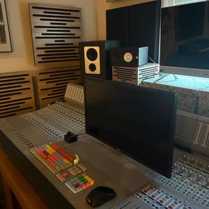Puede incluir: Un estudio de grabación profesional con una gran consola de mezclas, altavoces y paneles acústicos. La consola tiene botones coloridos y un monitor de ordenador. La sala está diseñada para la producción de audio.
