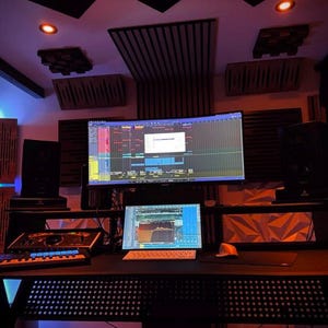 May include: A professional music studio setup with a large curved monitor displaying audio editing software. A smaller laptop screen shows a waveform. The desk includes a mixing console, keyboard, and soundproofing panels on the walls and ceiling.