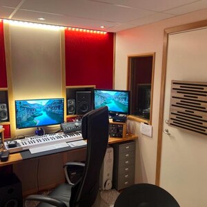 Puede incluir: Un estudio de grabación profesional con dos monitores de ordenador que muestran un paisaje fluvial. La sala cuenta con paneles insonorizantes, un teclado, altavoces y una silla de oficina negra. Las paredes son rojas, beige y blancas.