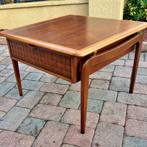 May include: A mid-century modern side table with a rectangular top and woven drawer. The table is made of wood with a warm brown finish. The legs are tapered and slightly curved, adding to its vintage appeal.