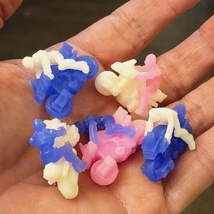 Könnte beinhalten: Eine Sammlung von Miniatur-Spielzeugmotorrädern in Blau, Rosa und Creme. Jedes Motorrad hat eine kleine Fahrerfigur. Die Spielzeuge werden in einer Hand gehalten, vor dunklem Hintergrund.