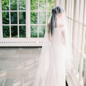 Puede incluir: Una novia con un vestido de novia blanco de hombros descubiertos y un velo largo y fluido. Est&aacute; de pie en una habitaci&oacute;n soleada con grandes ventanas.