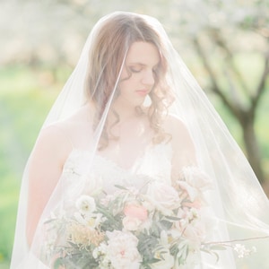 Puede incluir: Una novia con un vestido de novia blanco y velo sostiene un ramo de flores blancas y rosas. El ramo est&aacute; compuesto por rosas, peon&iacute;as y otras plantas verdes.