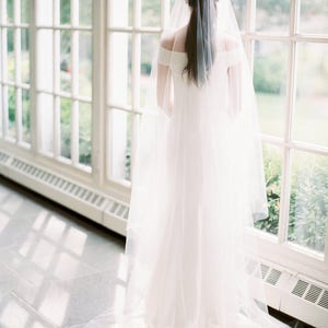 Puede incluir: Una novia con un vestido de novia blanco y un velo largo y fluido se encuentra frente a una gran ventana. El velo es transparente y delicado, y el vestido es sencillo y elegante. La novia mira por la ventana, y la luz de la ventana proyecta un suave resplandor sobre ella.