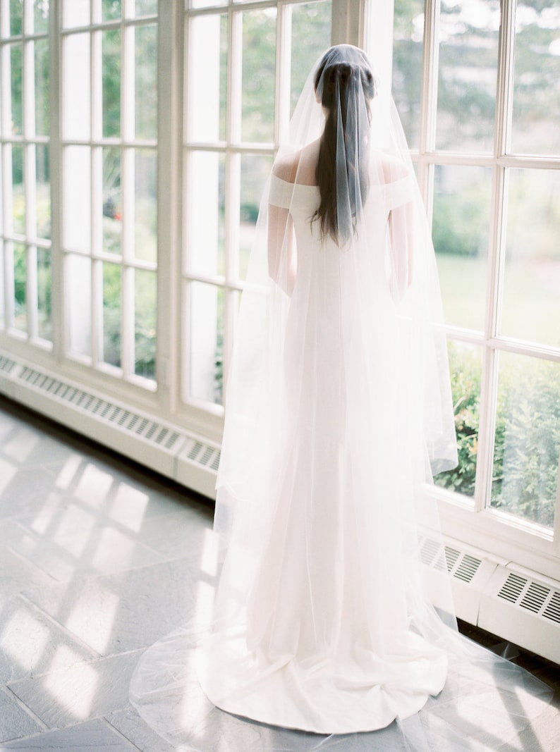Puede incluir: Una novia con un vestido de novia blanco y un velo largo y fluido se encuentra frente a una gran ventana. El velo es transparente y delicado, y el vestido es sencillo y elegante. La novia mira por la ventana, y la luz de la ventana proyecta un suave resplandor sobre ella.