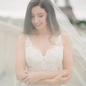 Puede incluir: Una novia con un vestido de novia de encaje blanco con un velo sobre su rostro. Ella sonr&iacute;e y mira hacia abajo. La Torre Eiffel est&aacute; en el fondo.