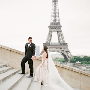 Puede incluir: Una novia y un novio tomados de la mano y subiendo una escalera con la Torre Eiffel de fondo.