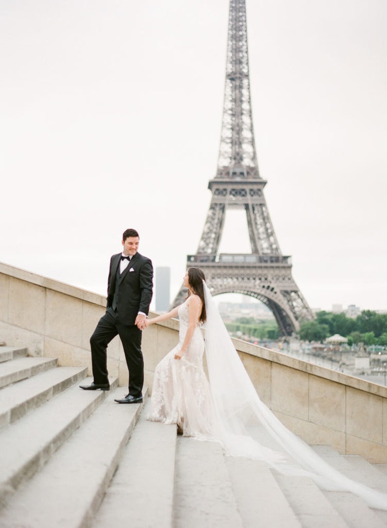 Puede incluir: Una novia y un novio tomados de la mano y subiendo una escalera con la Torre Eiffel de fondo.