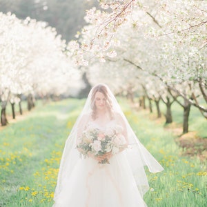 Puede incluir: Una novia con un vestido de novia blanco y velo se encuentra en un campo de hierba verde y flores amarillas. Lleva un ramo de flores blancas y rosas. El fondo es una hilera de &aacute;rboles de cerezo en flor.