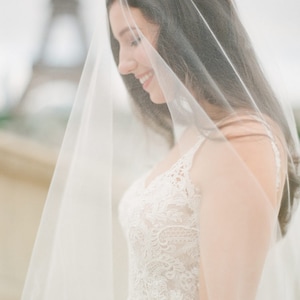 Puede incluir: Una novia con un vestido de novia de encaje blanco con un velo que le cubre la cara. La novia sonr&iacute;e y mira hacia abajo. La Torre Eiffel es visible en el fondo.