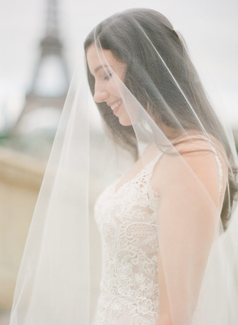 Puede incluir: Una novia con un vestido de novia de encaje blanco con un velo que le cubre la cara. La novia sonr&iacute;e y mira hacia abajo. La Torre Eiffel es visible en el fondo.