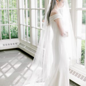 Puede incluir: Una novia con un vestido de novia blanco con un velo largo y una corona dorada. Est&aacute; de pie en una habitaci&oacute;n soleada con grandes ventanas.